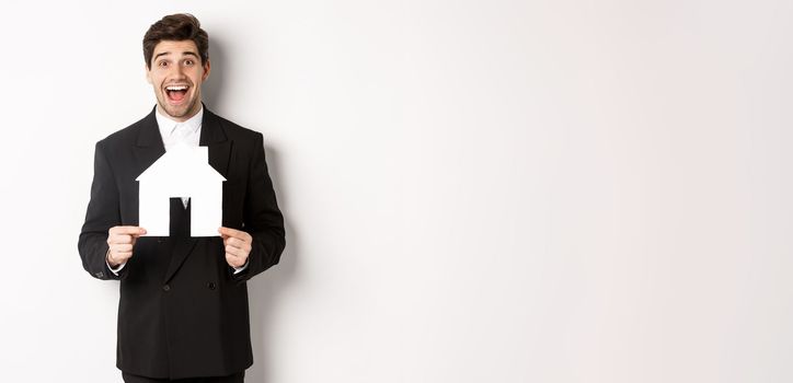 Image of handsome real estate agent in black suit showing home maket, and looking amazed, selling houses, standing against white background.