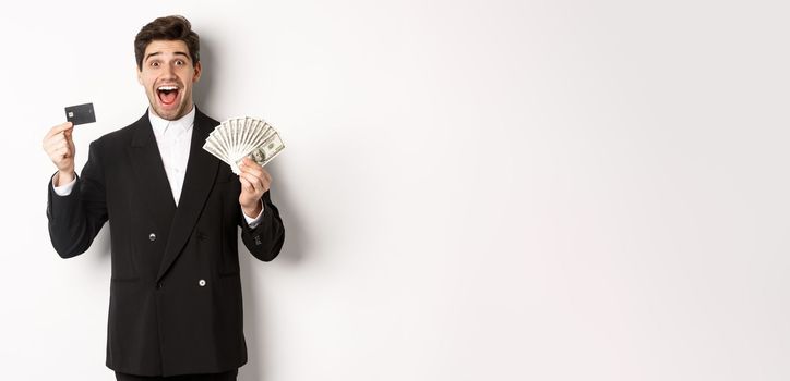 Image of handsome male entrepreneur in suit, showing credit card and money, smiling amazed, standing over white background.