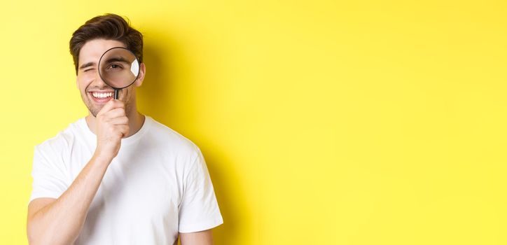 Close-up of young man looking through magnifying glass and smiling, searching something, standing over yellow background.