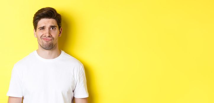 Close-up of dispelased young man in white t-shirt looking doubtful, grimacing unsatisfied, standing over yellow background.