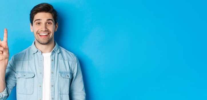 Close-up of handsome man smiling, showing fingers number two, standing over blue background.