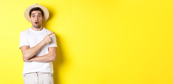 Surprised man in straw hat pointing finger right, showing promo banner, standing over yellow background. Copy space