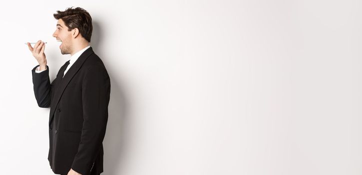 Profile shot of handsome businessman in black suit talking on speakerphone, smiling and looking happy, recording voice message, standing over white background.