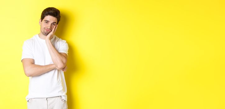 Bored and unamused man looking with indifference, standing in white clothes over yellow background.