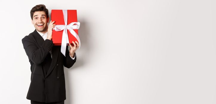 Concept of christmas holidays, celebration and lifestyle. Attractive man in black suit, holding new year gift and smiling, standing with a present over white background.