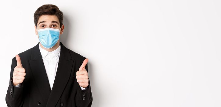 Concept of covid-19, business and social distancing. Close-up of happy businessman in black suit and medical mask, showing thumbs-up, making a compliment, white background.