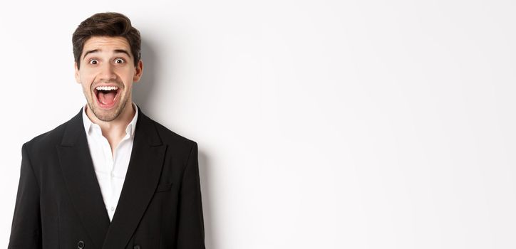 Close-up of attractive man in black suit, smiling amazed and looking at advertisement, standing over white background.