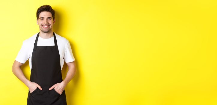 Happy barista in black apron looking at camera. Coffee shop owner wearing cafe uniform and smiling, standing over yellow background.