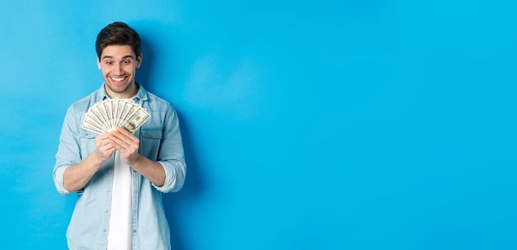 Excited successful man counting money, looking satisfied at cash and smiling, standing over blue background.