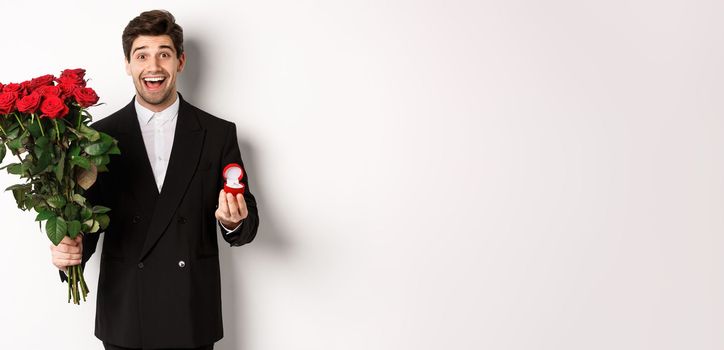 Handsome smiling man in black suit, holding roses and engagement ring, making a proposal to marry him, standing against white background.