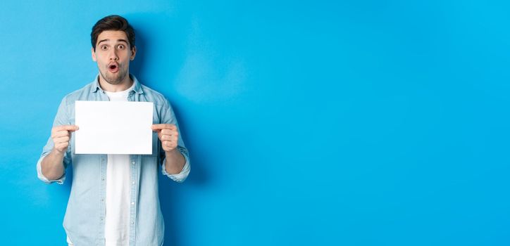 Surprised man gasping and looking impressed at camera, showing blank piece of paper for your sign, standing over blue background.