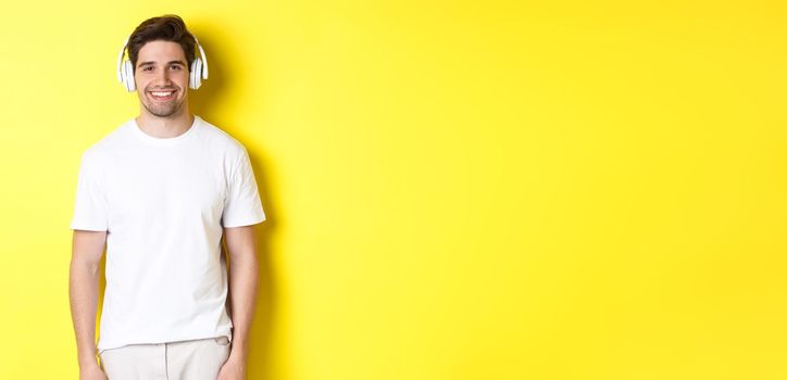 Young handsome man listening music in headphones, wearing earphones and smiling, standing over yellow background.