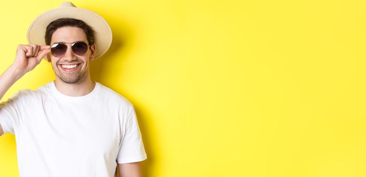 Concept of tourism and vacation. Close-up of handsome man tourist looking happy, wearing sunglasses and summer hat, standing over yellow background.