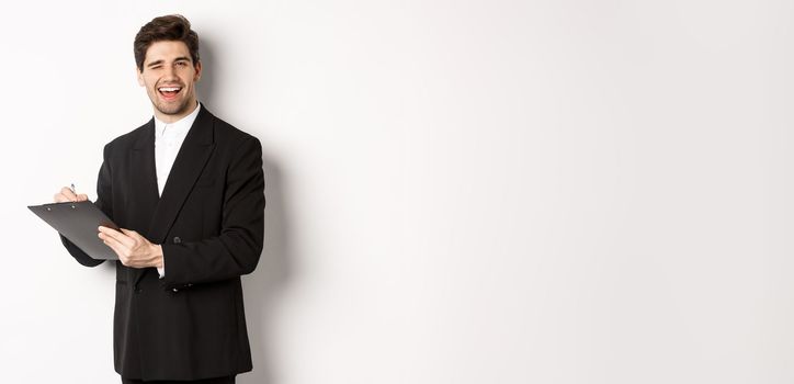 Image of handsome, successful boss in black suit, winking and smiling while signing documents, standing against white background.