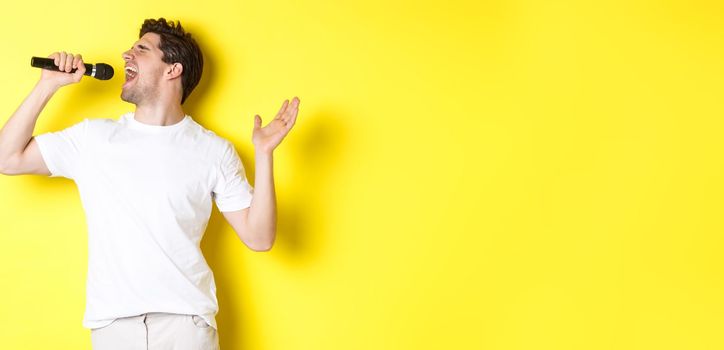 Young man singer holding microphone, reaching high note and singing karaoke, standing over yellow background.