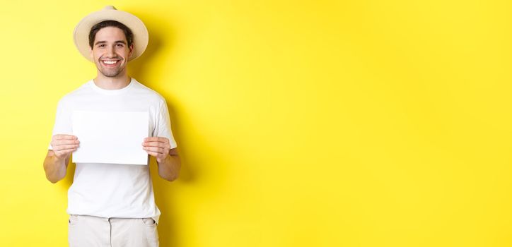 Handsome young male tourist in summer hat smiling, holding blank piece of paper for your sign, standing over yellow background.