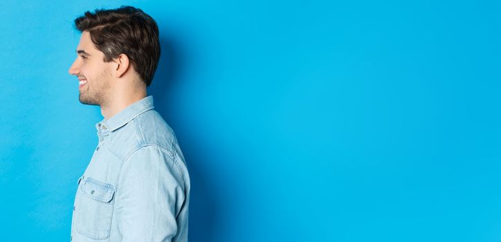 Profile of handsome young man looking left, smiling happy, standing over blue background.