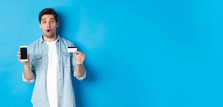 Amazed young man showing mobile cell phone screen and credit card, shop online, standing against blue background.