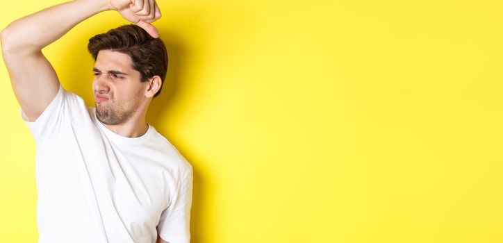 Man in sweat smelling his armpit, standing in white t-shirt and grimacing from stinky clothes.