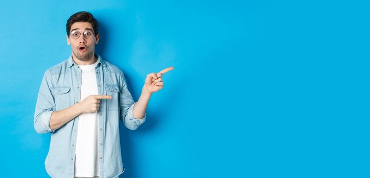 Surprised man in glasses pointing right at copy space, showing promo offer on blue background, standing over blue background.