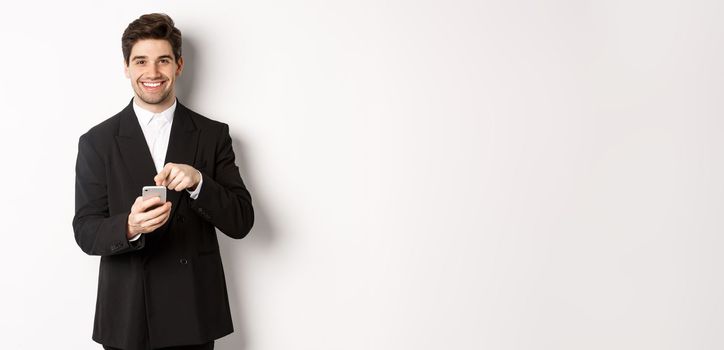 Image of handsome businessman in suit showing you app on smartphone, pointing finger at mobile screen and smiling, standing over white background.