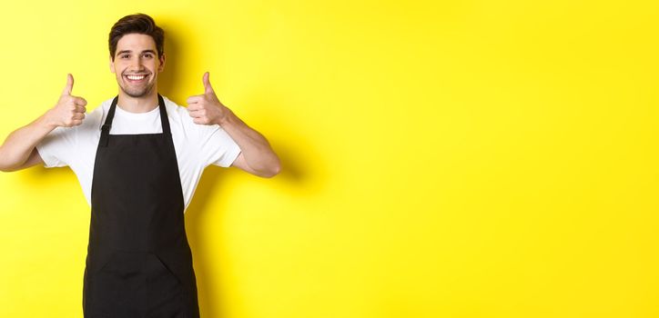 Smiling seller in black apron showing thumbs up, approve or like something, recommending cafe or store, yellow background.