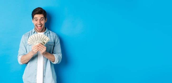 Excited successful man counting money, looking satisfied at cash and smiling, standing over blue background.