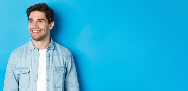 Young modern guy with beard, looking left and smiling pleased at copy space, standing over blue background.