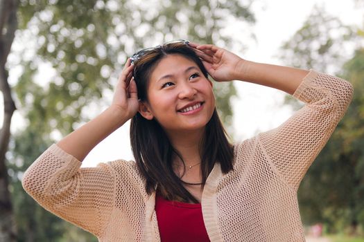 portrait of a cheerful asian woman putting on her sunglasses on her head smiling in the park, concept of youth and beauty