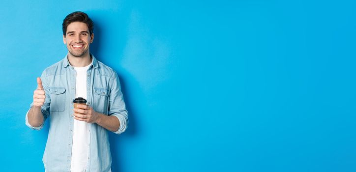 Handsome young man showing thumb-up and drinking coffee, recommending cafe takeaway, standing over blue background.