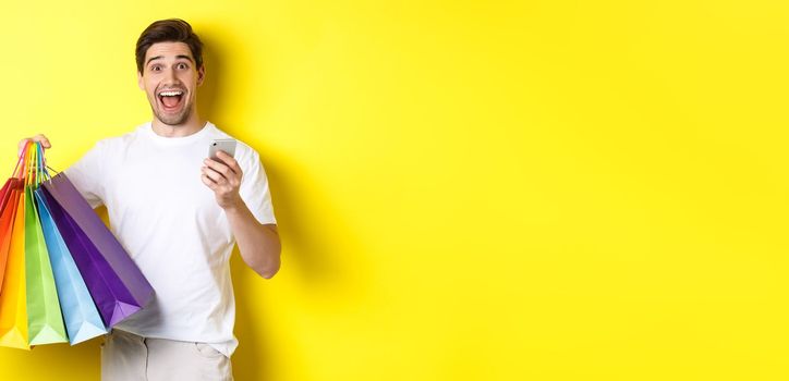 Image of happy man receive cashback for purchase, holding smartphone and shopping bags, smiling excited, standing over yellow background.