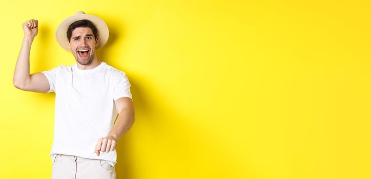 Concept of tourism and summer. Young man traveller showing rodeo gesture, standing in straw hat and white clothes, standing over yellow background.