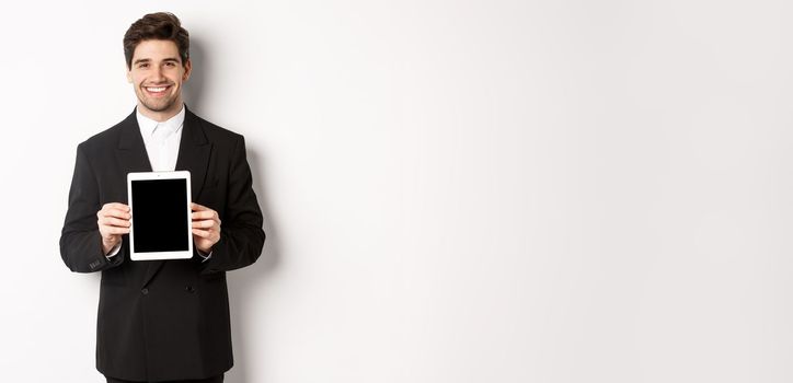 Portrait of handsome businessman in trendy suit, showing digital tablet screen and smiling, standing against white background.