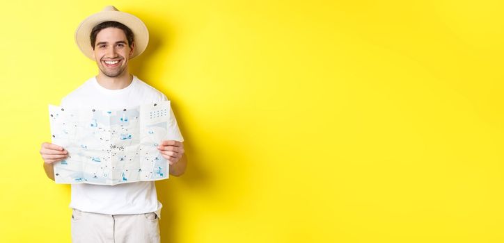 Travelling, vacation and tourism concept. Smiling young man going on trip, holding road map and smiling, standing over yellow background.