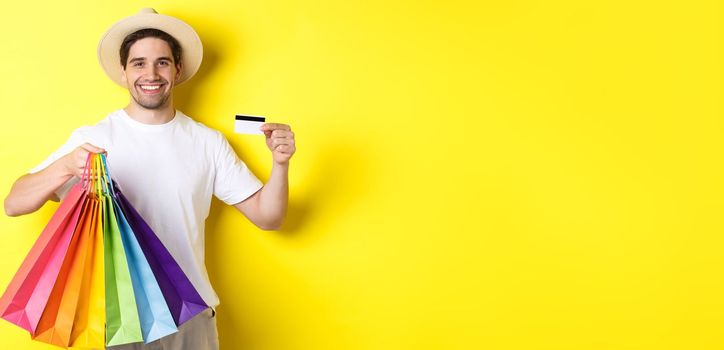 Happy attractive guy showing shopping bags and credit card, concept of banking and easy payment, standing over yellow background.