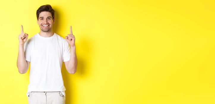 Handsome guy in white t-shirt pointing fingers up, showing shopping offers, standing over yellow background.