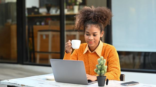 Pleased female freelancer drinking coffee and working online on laptop while sitting outdoor.