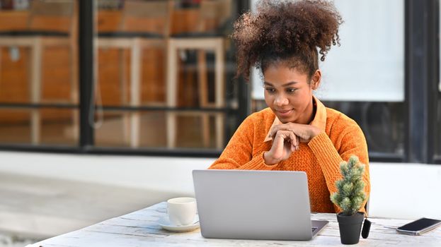 Focused African American woman watching online webinar on laptop computer while sitting at outdoor cafe.