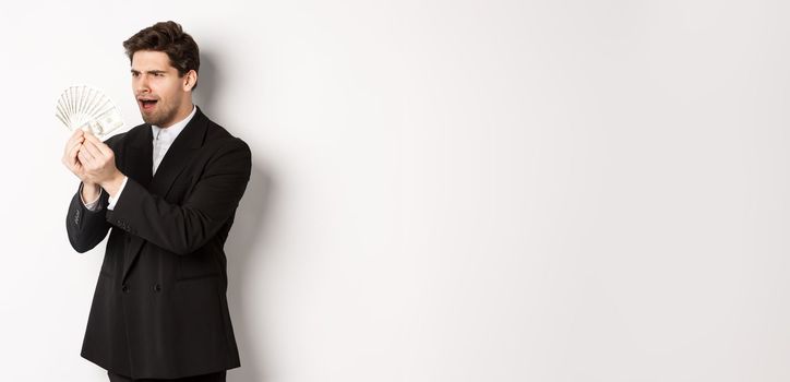 Image of confused businessman looking at fake money, standing over white background in black suit.