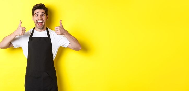 Cheerful man barista in black apron showing thumbs-up, recommending cafe or restaurant, standing against yellow background.