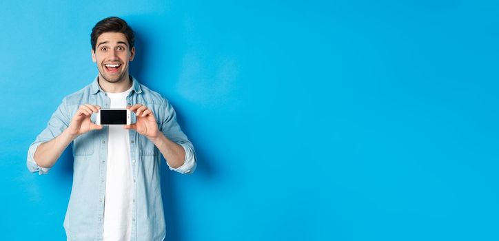 Amazed smiling man showing smartphone screen, internet promo offer, standing against blue background.