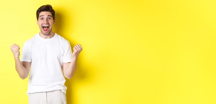 Excited and lucky man winning, clench fists and looking happy, triumphing and celebrating, standing over yellow background.