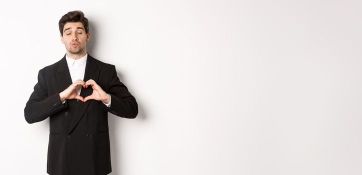 Image of handsome groom in black suit, showing heart sign, close eyes and pucker lips, waiting for kiss, standing against white background.