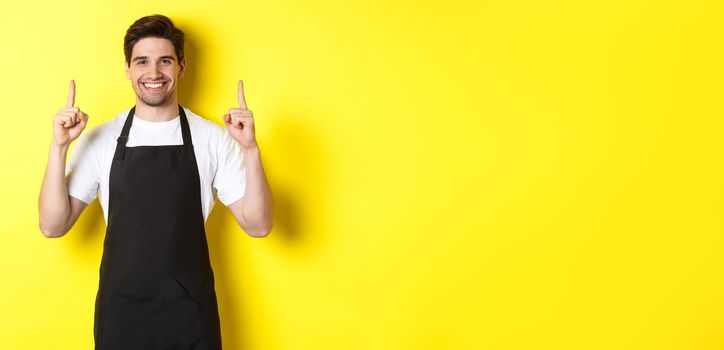 Handsome barista showing advertisement, coffee shop promo offer, pointing fingers up, standing against yellow background.