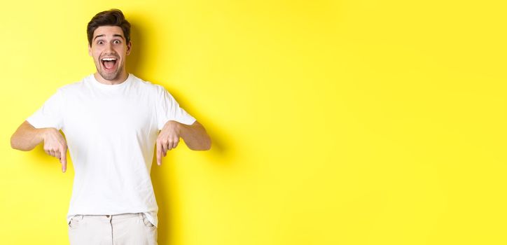 Amazed handsome man pointing fingers down, showing banner with excitement, standing over yellow background.