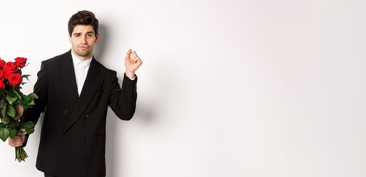 Image of elegant and sassy man in black suit, looking confident and holding bouquet of red roses, going on a romantic date, standing against white background.