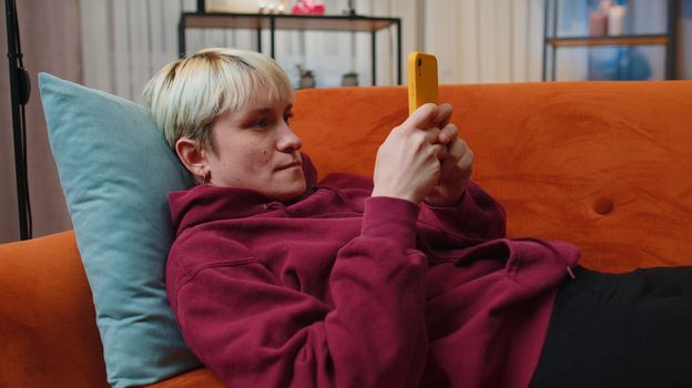 Young woman with short blonde hair lying on sofa uses mobile phone smiles at night evening home. Girl texting share messages content on smartphone social media applications online watching relax movie