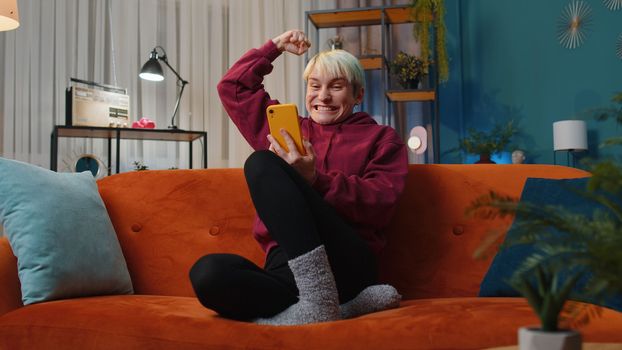 Happy woman with short hair use mobile smartphone typing browsing say Wow yes found out great big win good news celebrate lottery jackpot doing winner gesture. Girl at home in room sitting on sofa