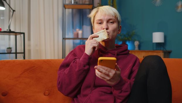 Young woman with short hair using credit bank card and smartphone while transferring money, purchases online shopping, order food delivery at home apartment indoors. Girl in evening room on couch