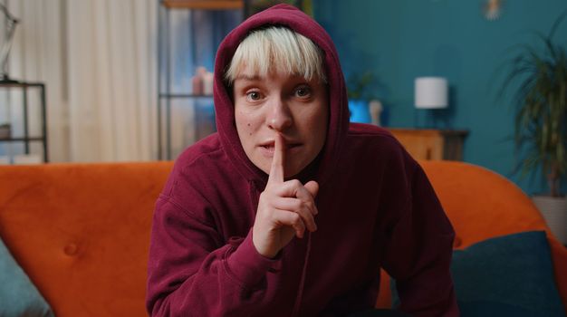 Shh be quiet, hush please. Young woman with short blonde hair presses index finger to lips makes silence hush gesture sign do not tells gossip secret. Girl at modern home evening room sitting on couch
